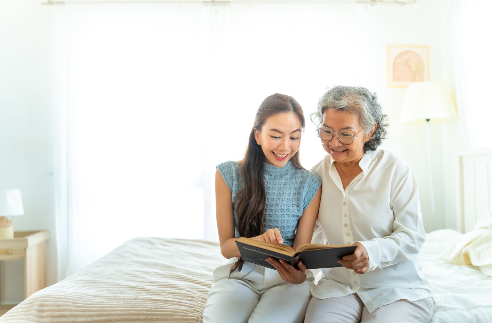 A grandchild and their grandparent in a bright sunlit bedroom reading a large hardcover book together.