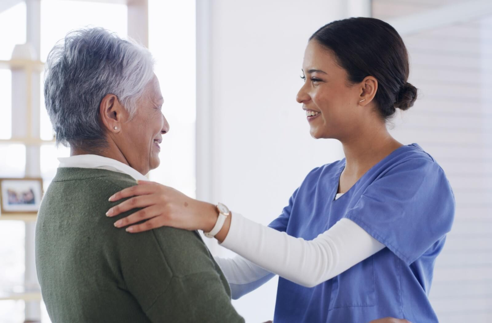 Caregiver in scrubs smiling while supporting a senior woman during assisted living care
