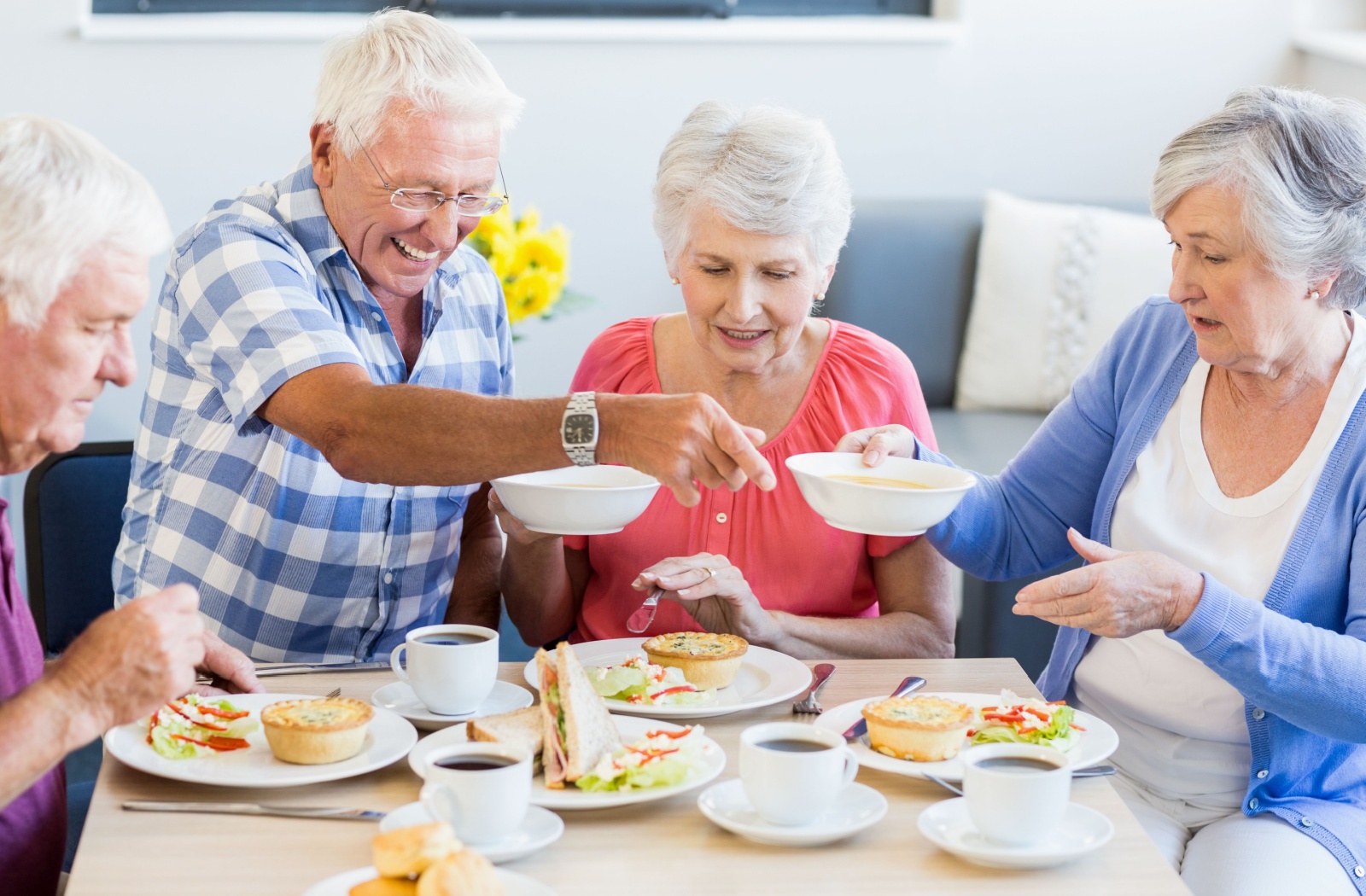 A group of older adults sit around a dining table in a well-lit senior living home and pass dishes back and forth during a shared meal