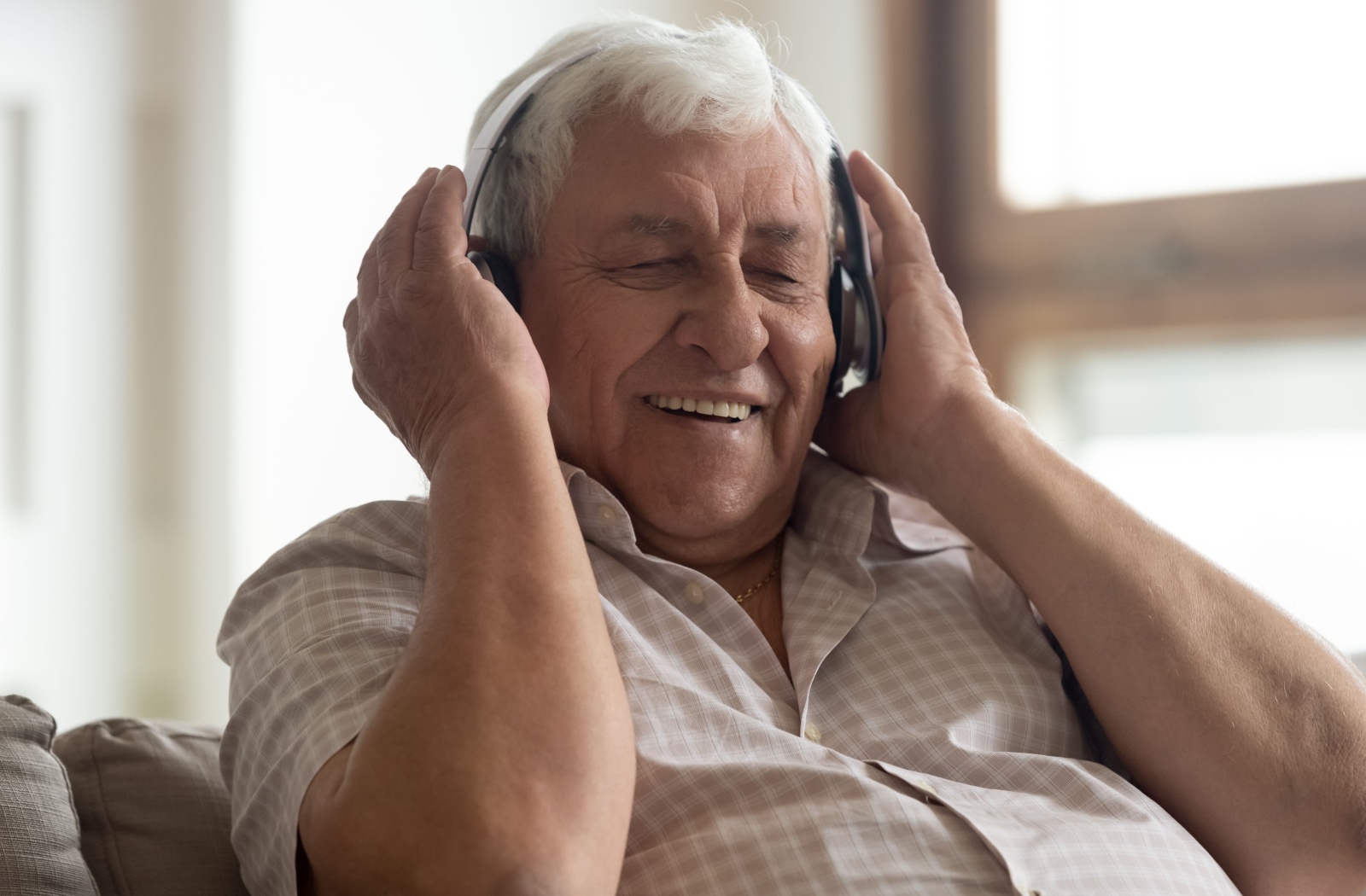 A senior sits on the couch, smiling and wearing headphones while listening to music.