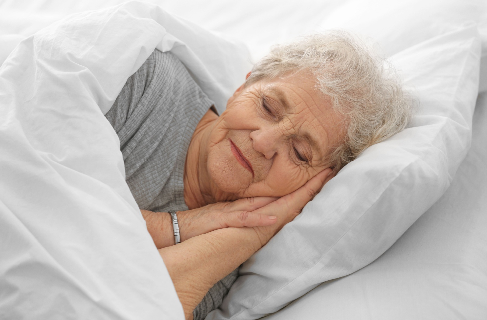 An older adult woman with grey hair sleeping alone on a bed with her eyes closed.