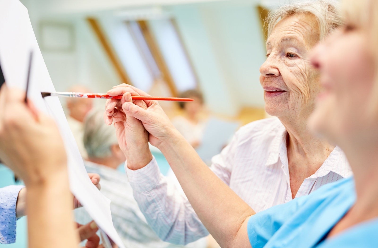 A staff member helping an older adult in senior living hold a paintbrush to white paper.
