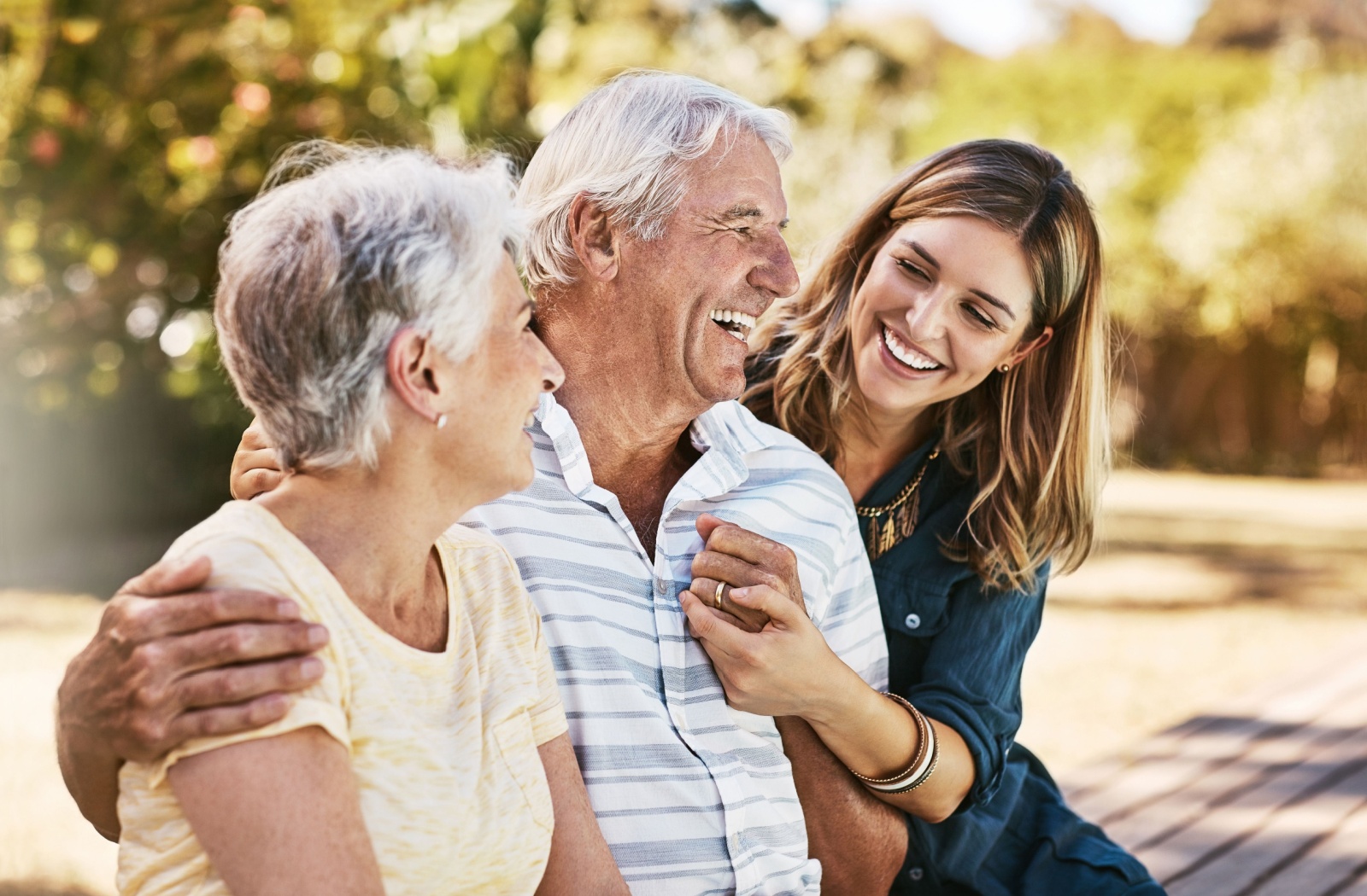 A smiling adult woman hugs her smiling senior parents.