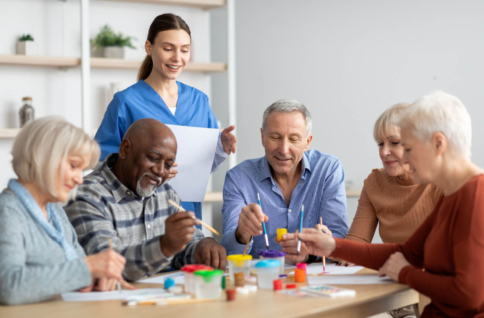 A caregiver smiling while checking in on a group of 5 happy residents during arts and crafts.