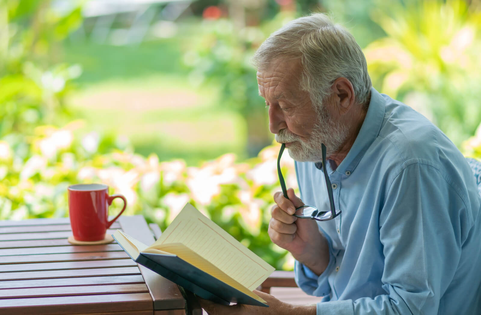 An older adult with dementia reading at a picnic table outside, chewing on their glasses arm thoughtfully.