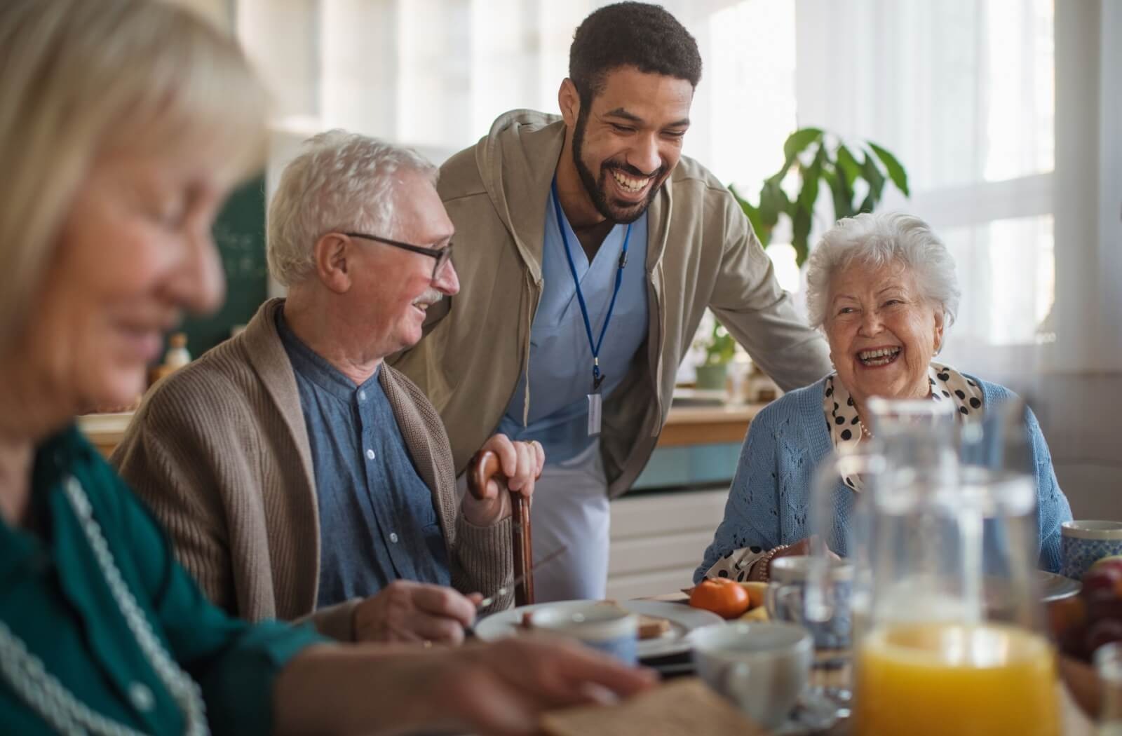 A laughing caregiver checks in on 3 happy residents after a communal breakfast in assisted living